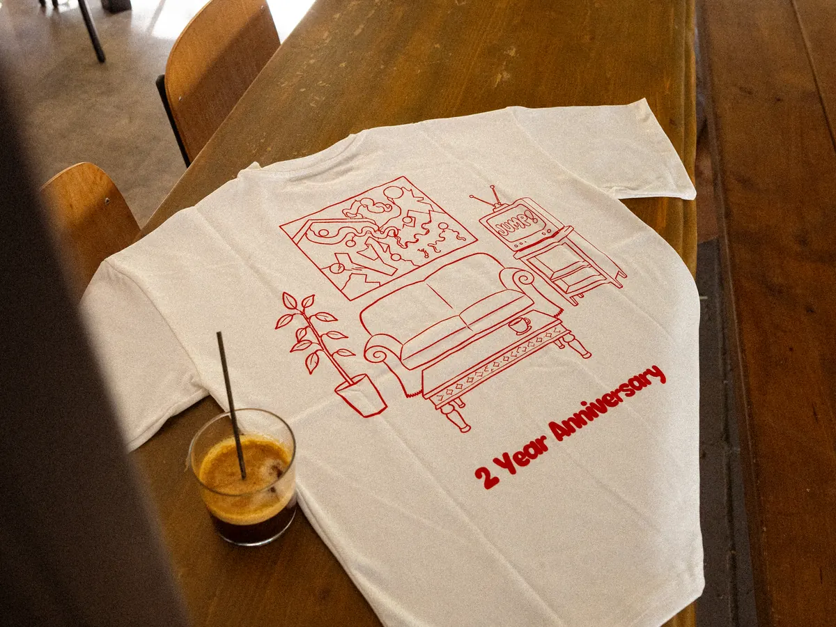 The anniversary shirt laid flat on a table, back print showing a line drawing of the room and “2 Year Anniversary”, next to an iced coffee