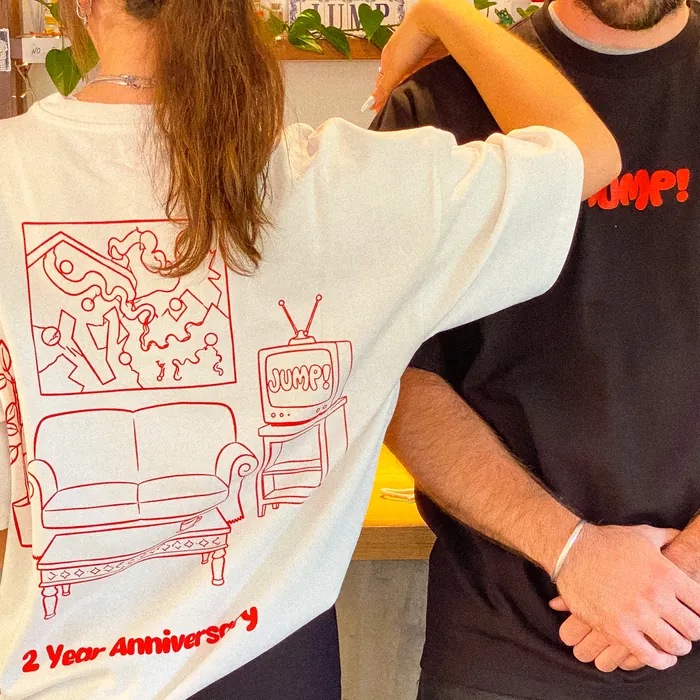 The white shirt from behind, the full back print in red line — a sofa, a painting and a TV reading Jump! — next to someone in the black shirt from the front