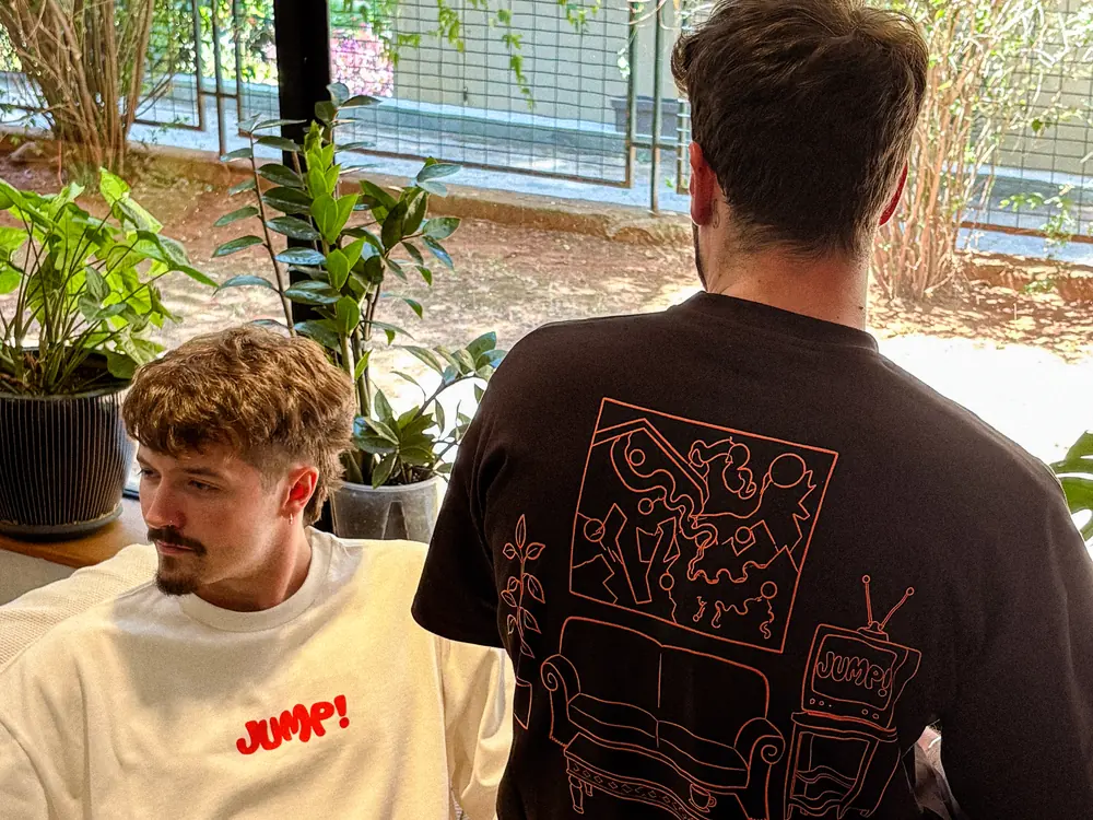 Two people on a sofa by the window in the shirts: one in white from the front, Jump! across the chest in red; one in black from behind, showing the back print of the room and “2 Year Anniversary”